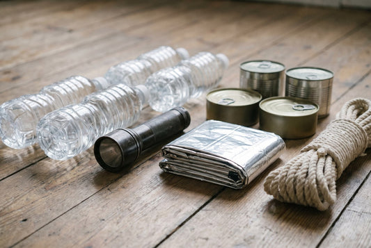 Emergency supplies laid out on a floor: sealed water bottles, a torch, a foil blanket, tinned food and cord
