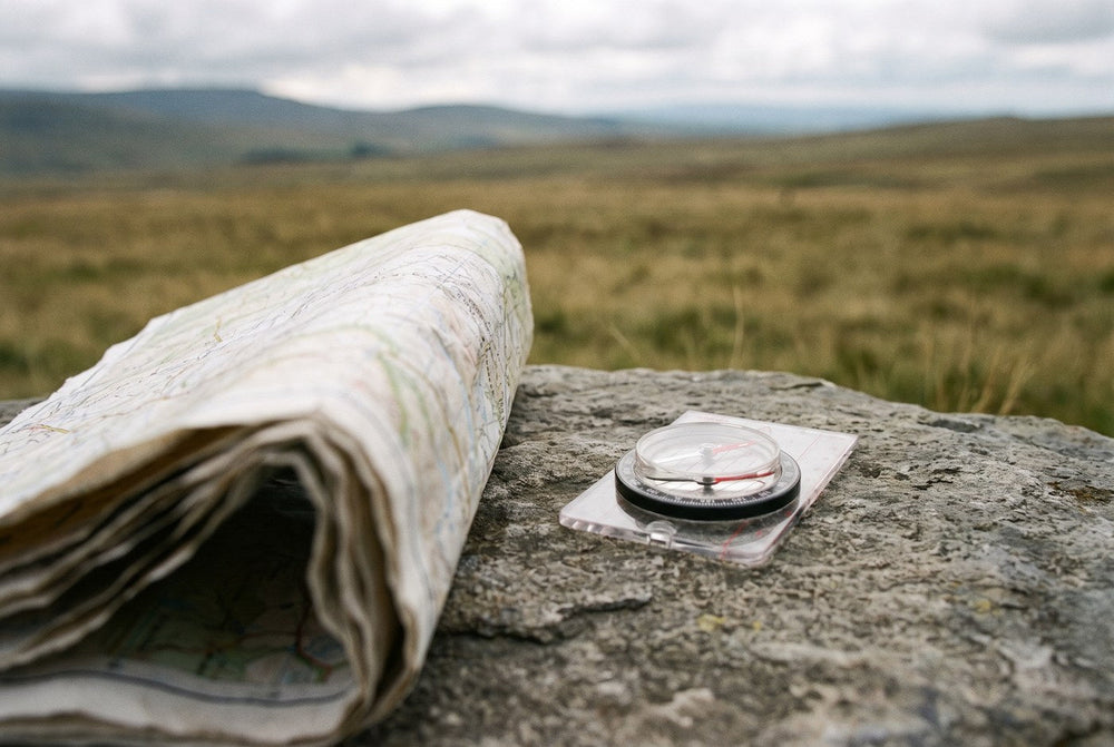 A folded paper map and a baseplate compass resting on a flat rock in open country